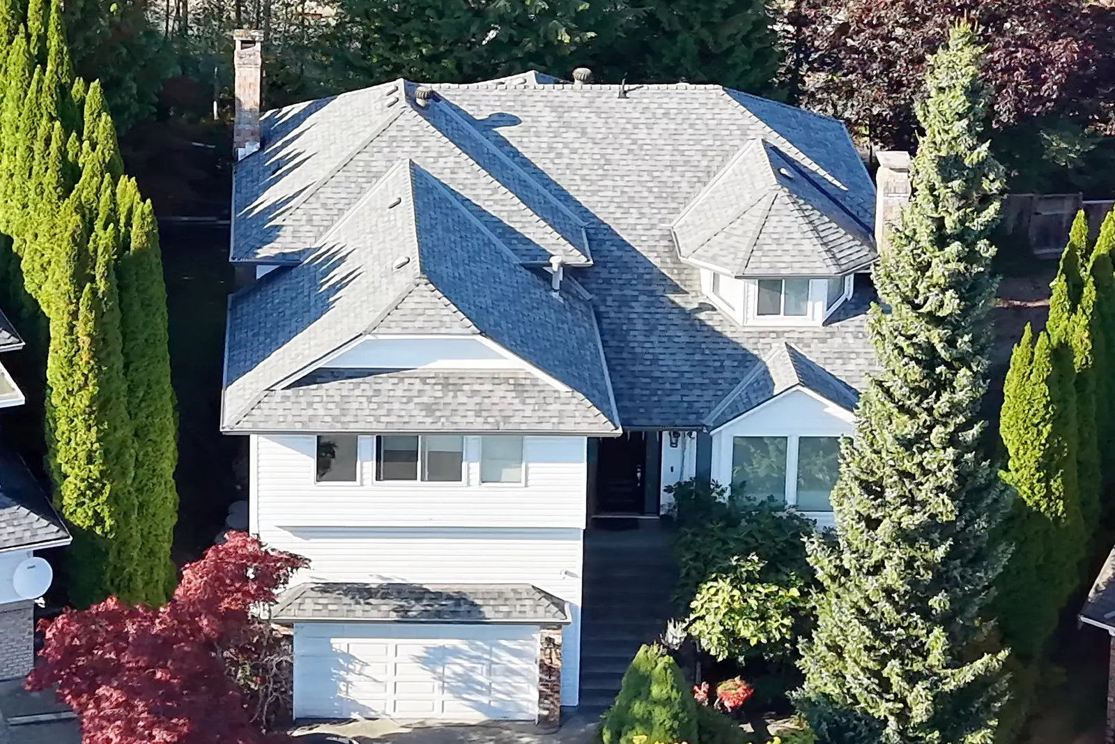 Front of a white two-storey house with a grey shingle roof and a double garage, set among tall evergreens and a red-leafed tree
